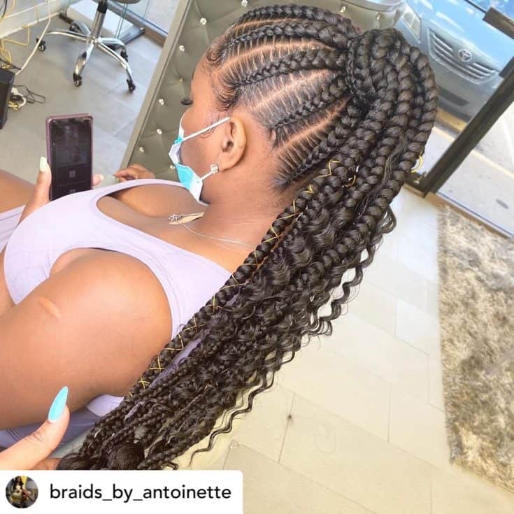 A person with intricately braided hair is sitting and holding a phone, partially turning towards the camera.