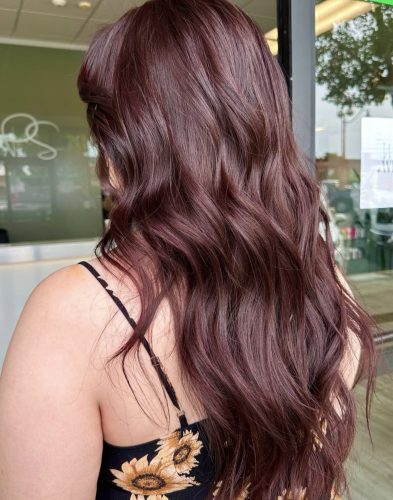 A woman with long, wavy dark burgundy hair standing indoors near a window. Her hair has a rich plum-red tone with soft, glossy waves. She is wearing a black dress with yellow sunflower prints, and the photo is taken from behind to show the color and texture of her hair.